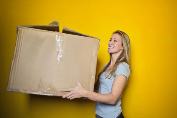 Blonde woman carrying large cardboard box against yellow background - visit rsd-storage.com.